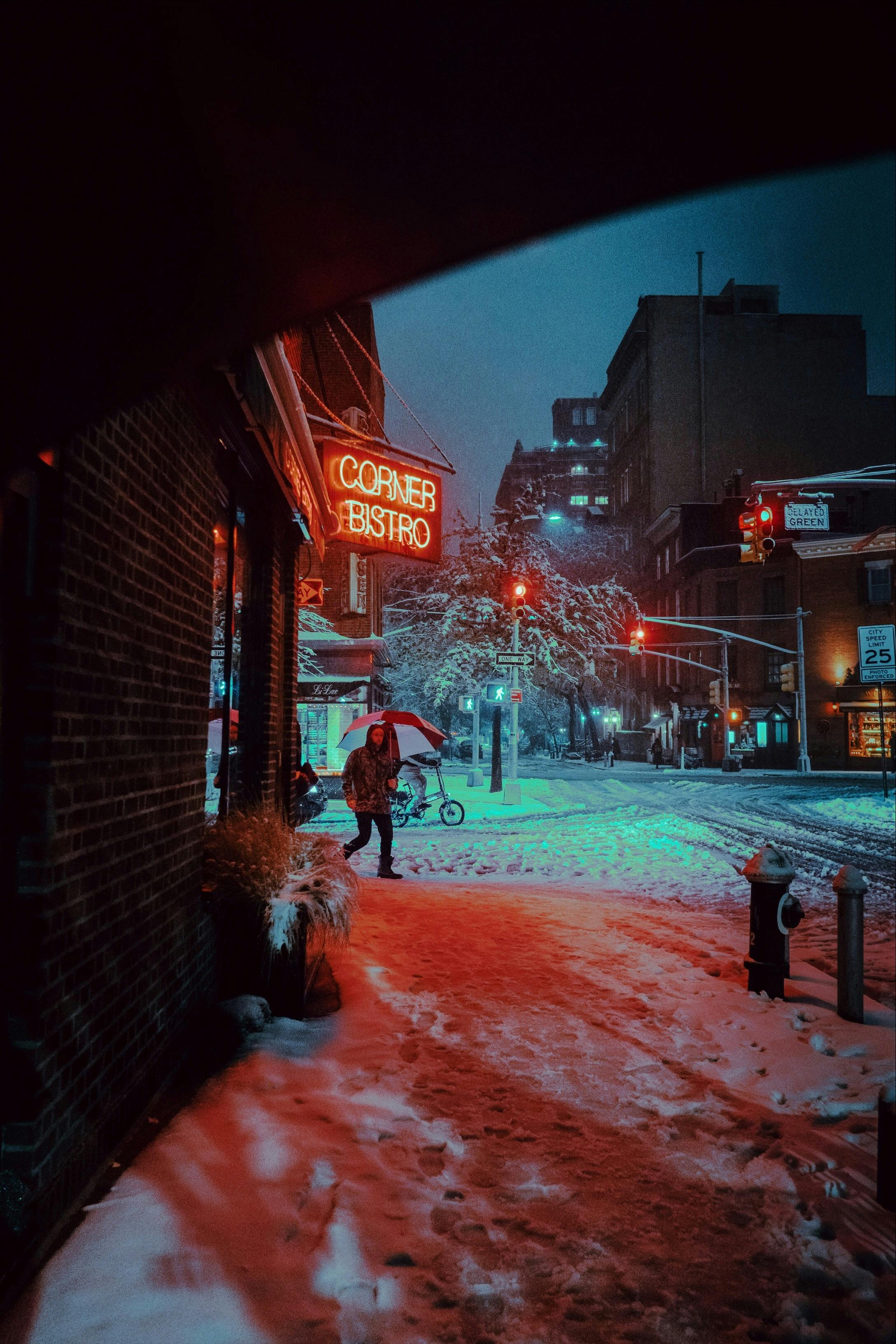 A snowy night in New York City featuring the "Corner Bistro" sign, with a person walking through the red and blue glow of city lights on the snow.