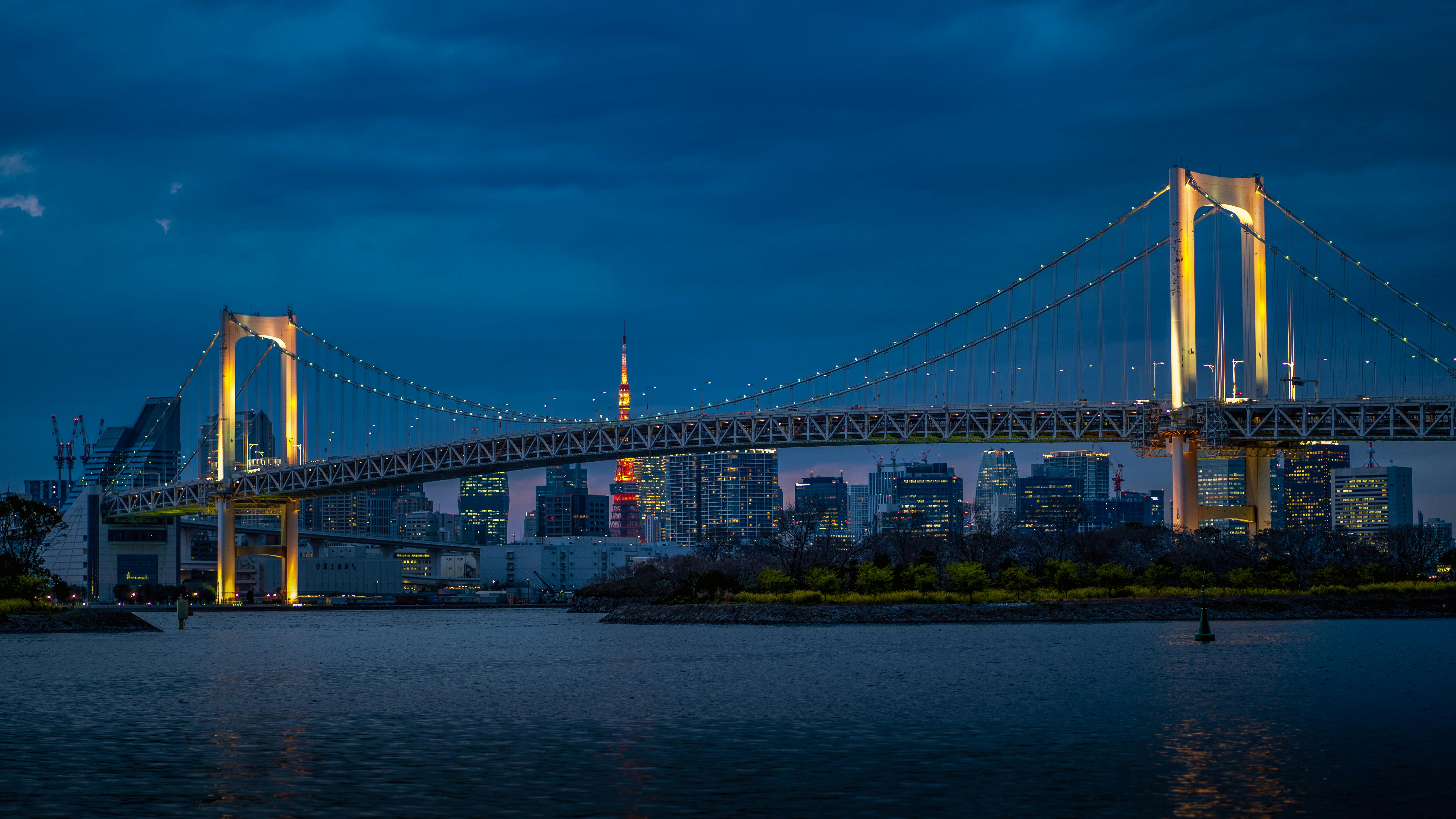 The Rainbow Bridge in Tokyo at twilight, with the city skyline and Tokyo Tower glowing in the distance across the water.