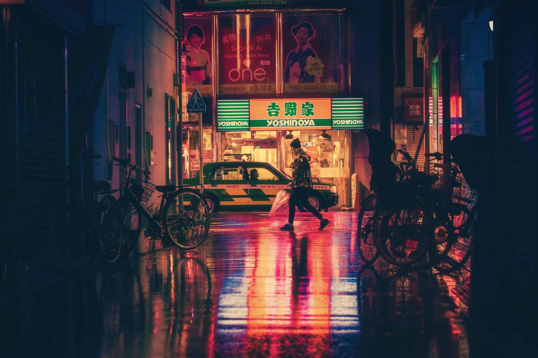A moody, rain-slicked Tokyo street at night with neon signs for Yoshinoya reflecting in the puddles as a person walks by.