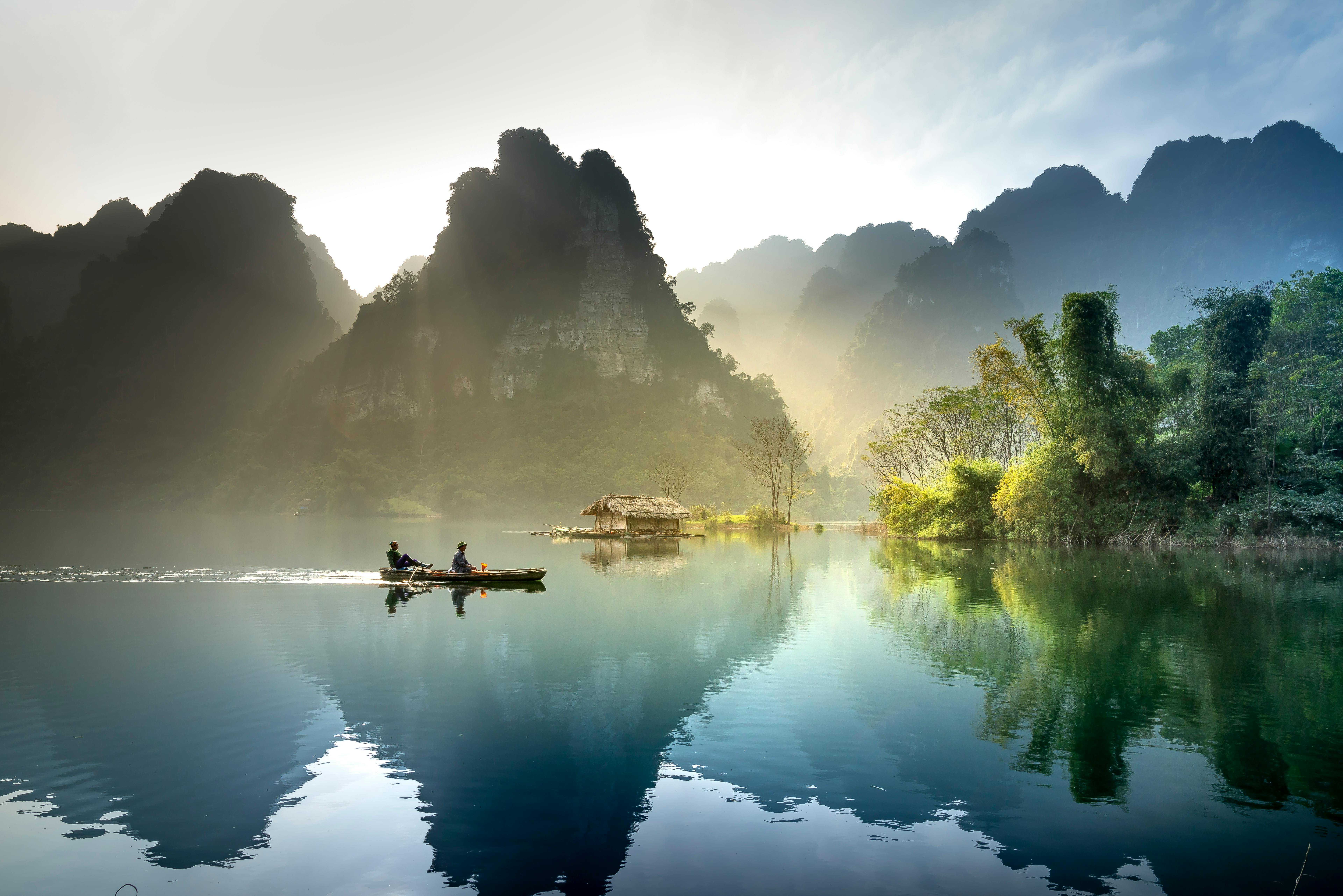 A misty morning on a Southeast Asian river, with tall limestone karsts in the background and a small wooden boat with two people in the foreground.