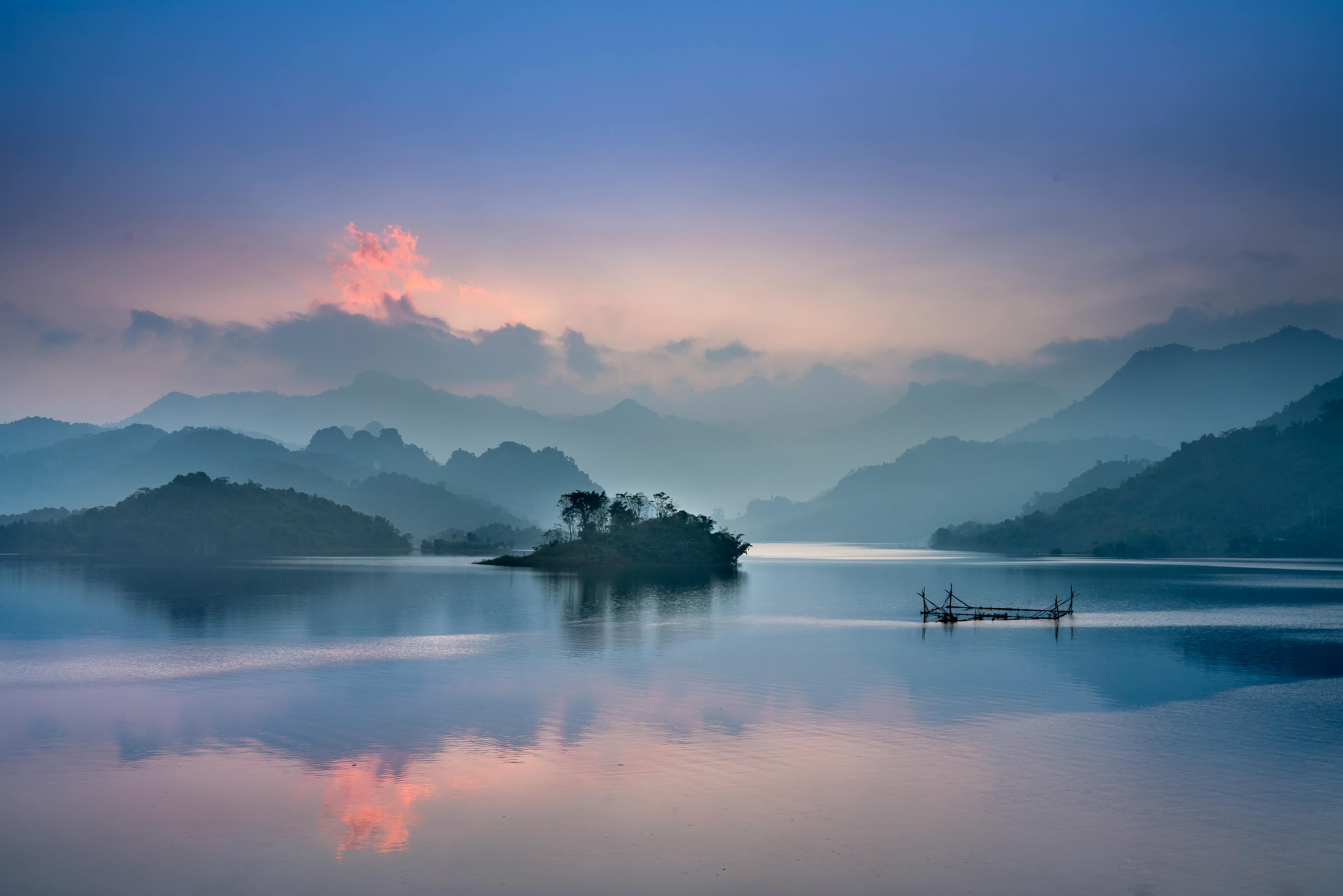 A calm, blue-hued lake at dawn, with small islands and distant mountains partially obscured by soft morning mist.