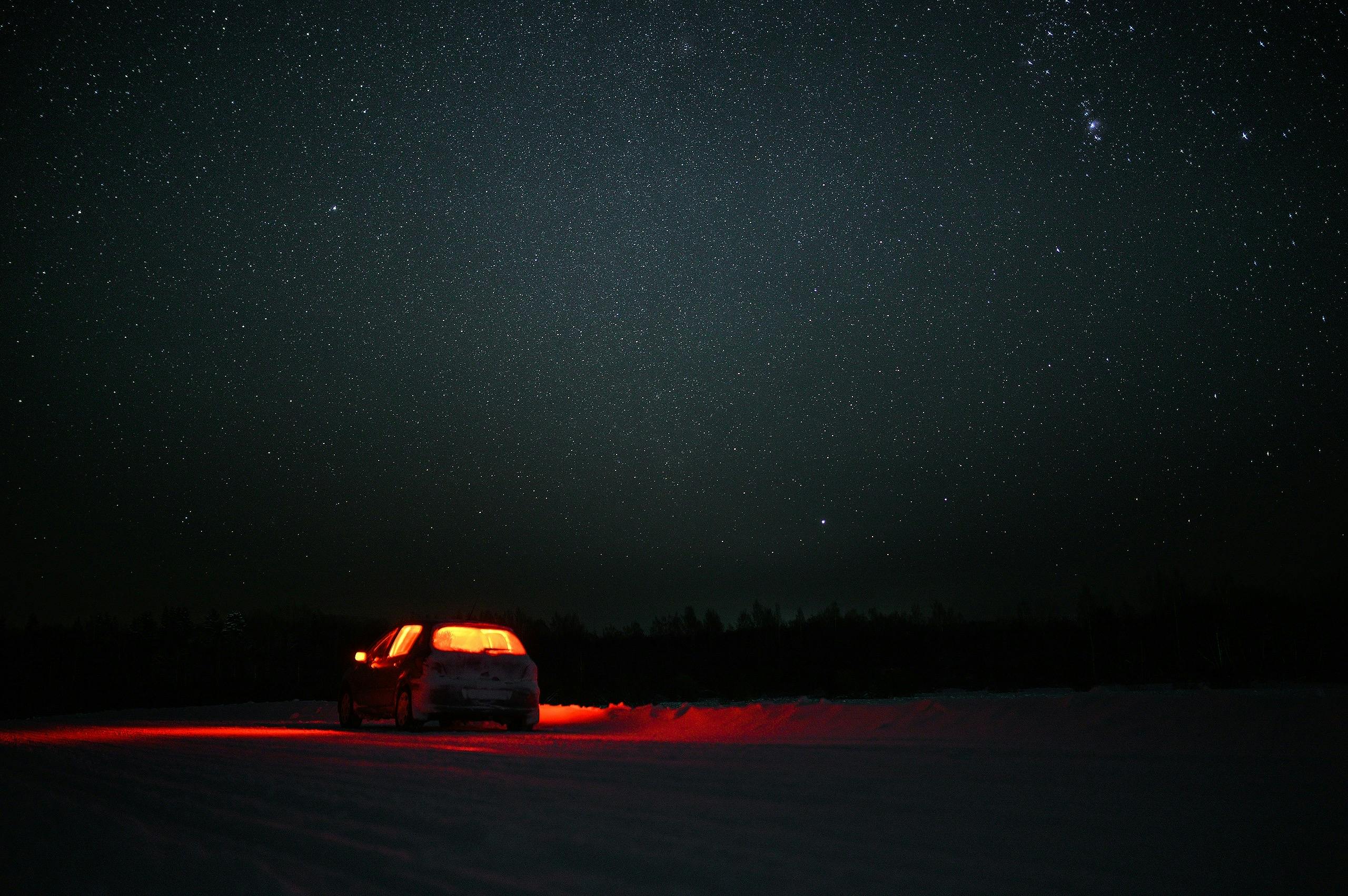 A car parked on a dark, open field at night, its red taillights glowing under a vast, star-filled sky.