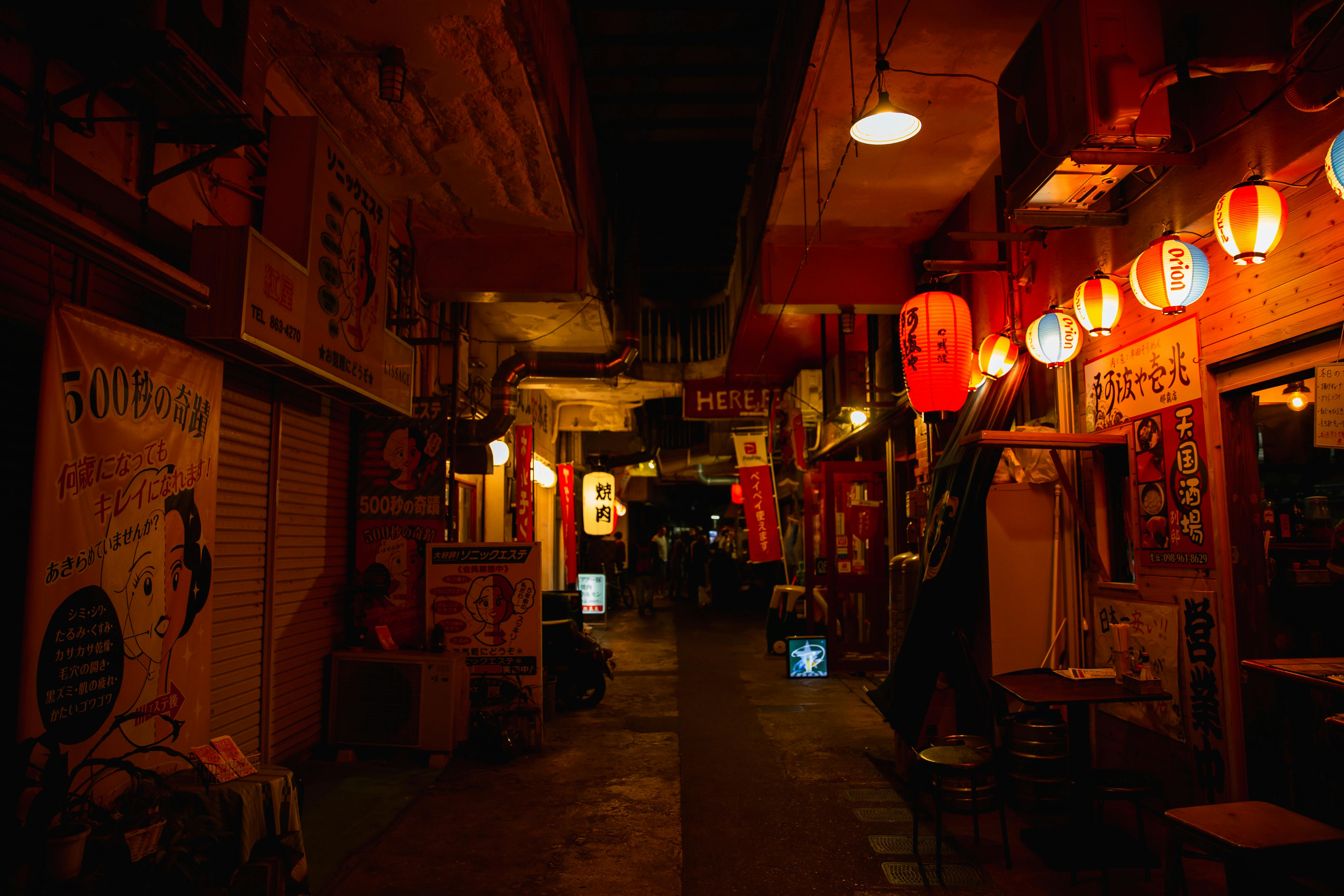 A narrow, dimly lit Japanese alleyway at night, lined with traditional wooden storefronts and glowing red paper lanterns.
