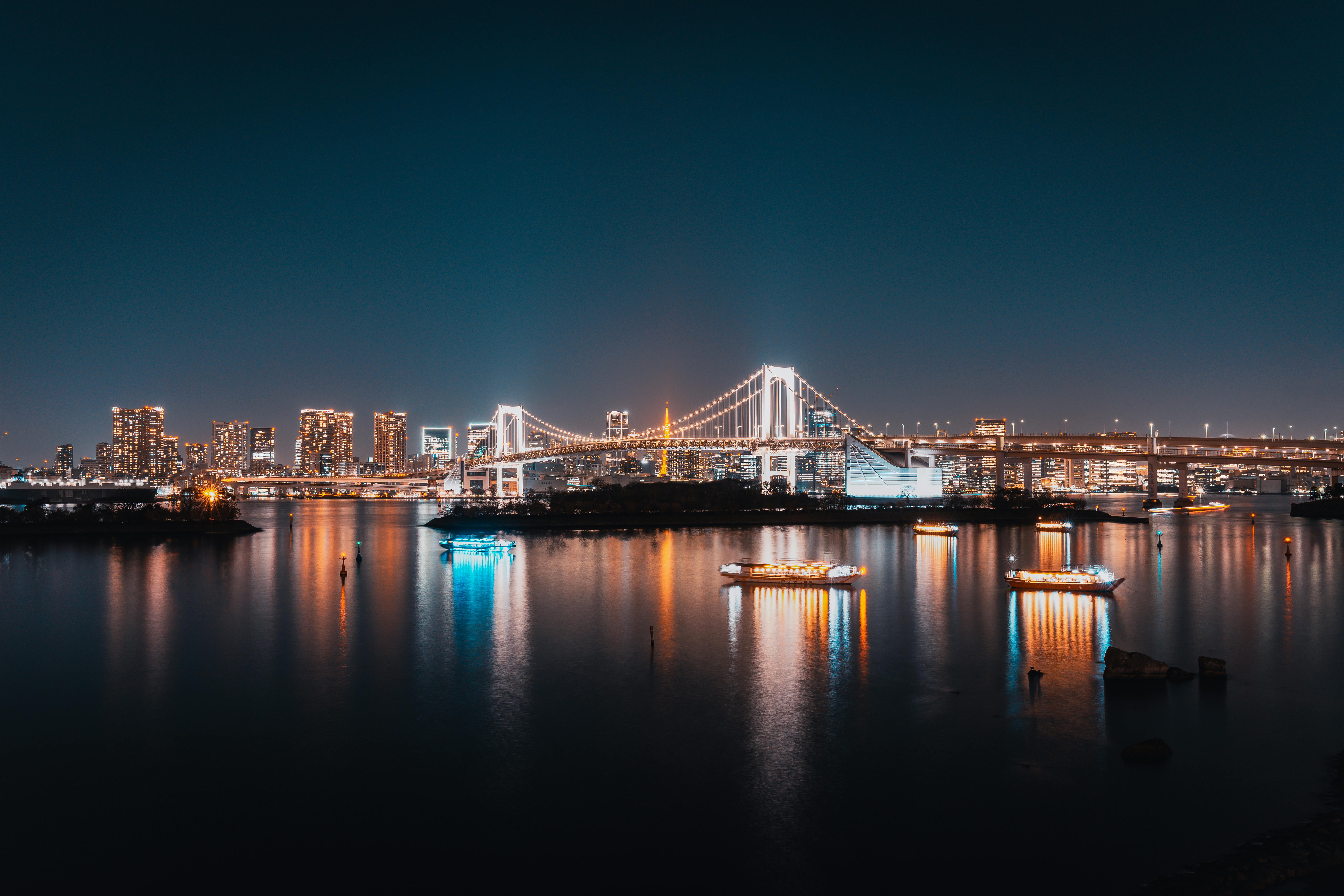 The Rainbow Bridge in Tokyo at night, illuminated in white light and reflecting perfectly on the dark water of the bay.