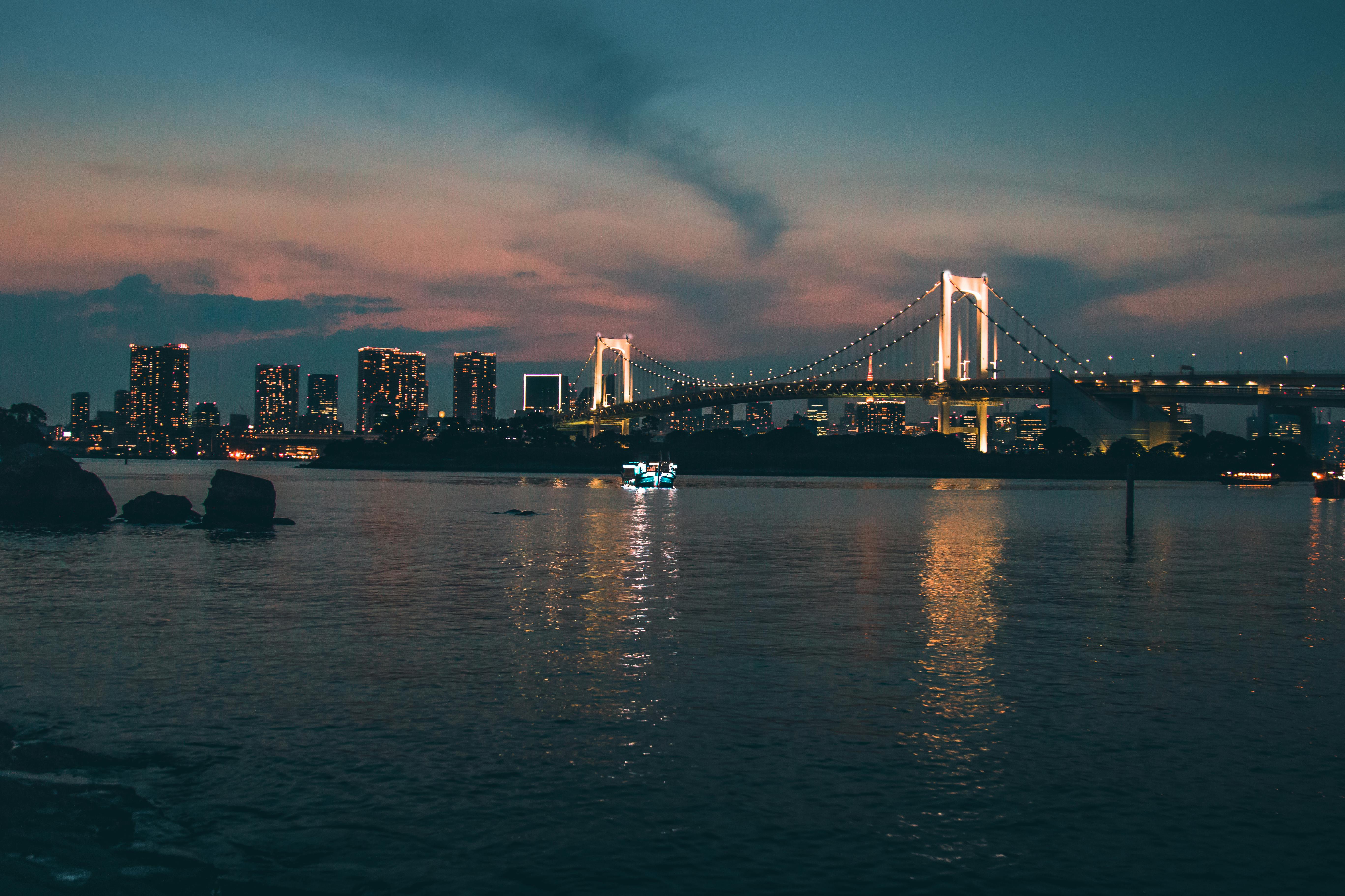A wide view of the Rainbow Bridge in Tokyo at dusk, with the city skyline illuminated and glowing lights reflecting on the calm water of the bay.