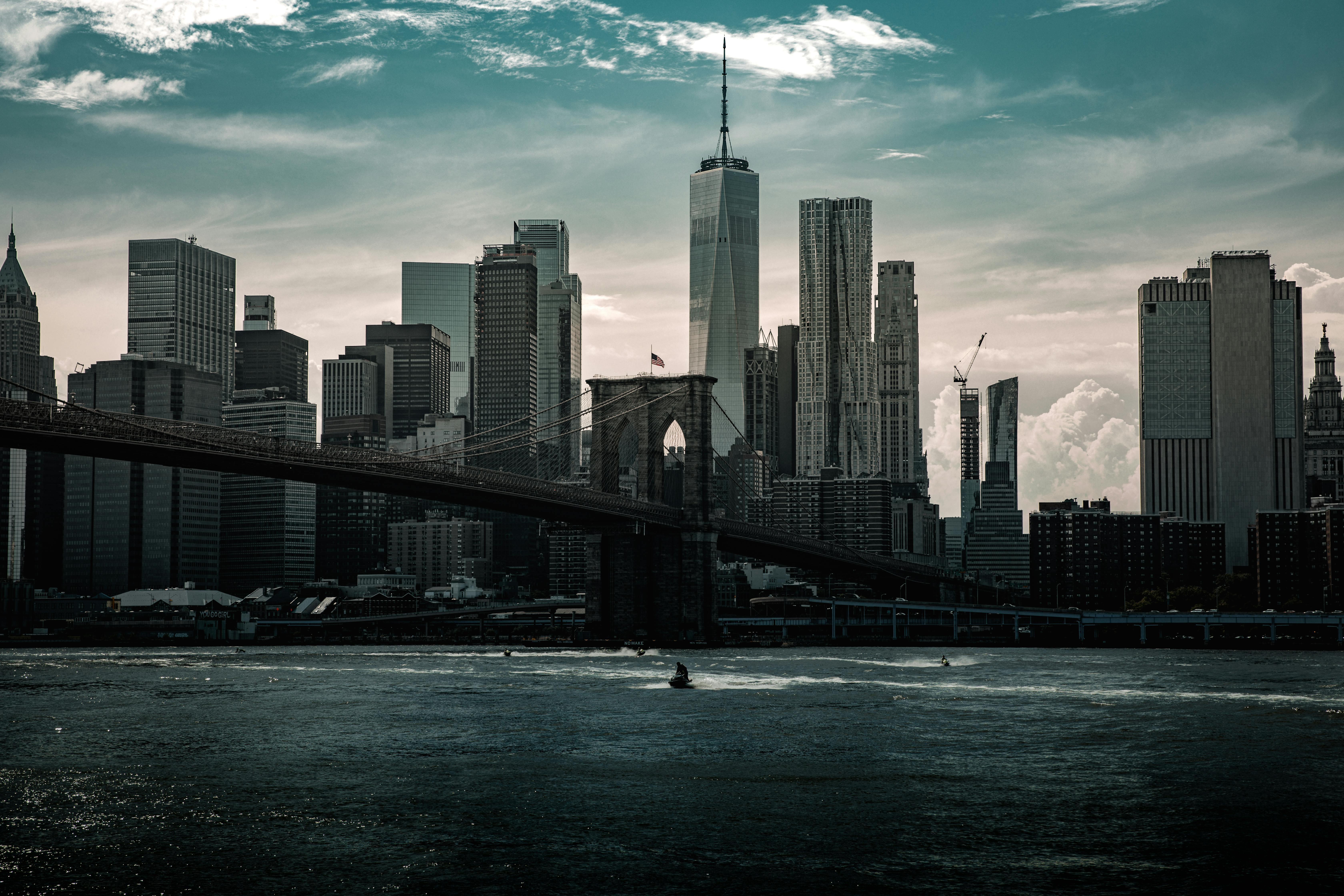 A moody, desaturated view of the Lower Manhattan skyline and One World Trade Center behind the dark silhouette of the Brooklyn Bridge.