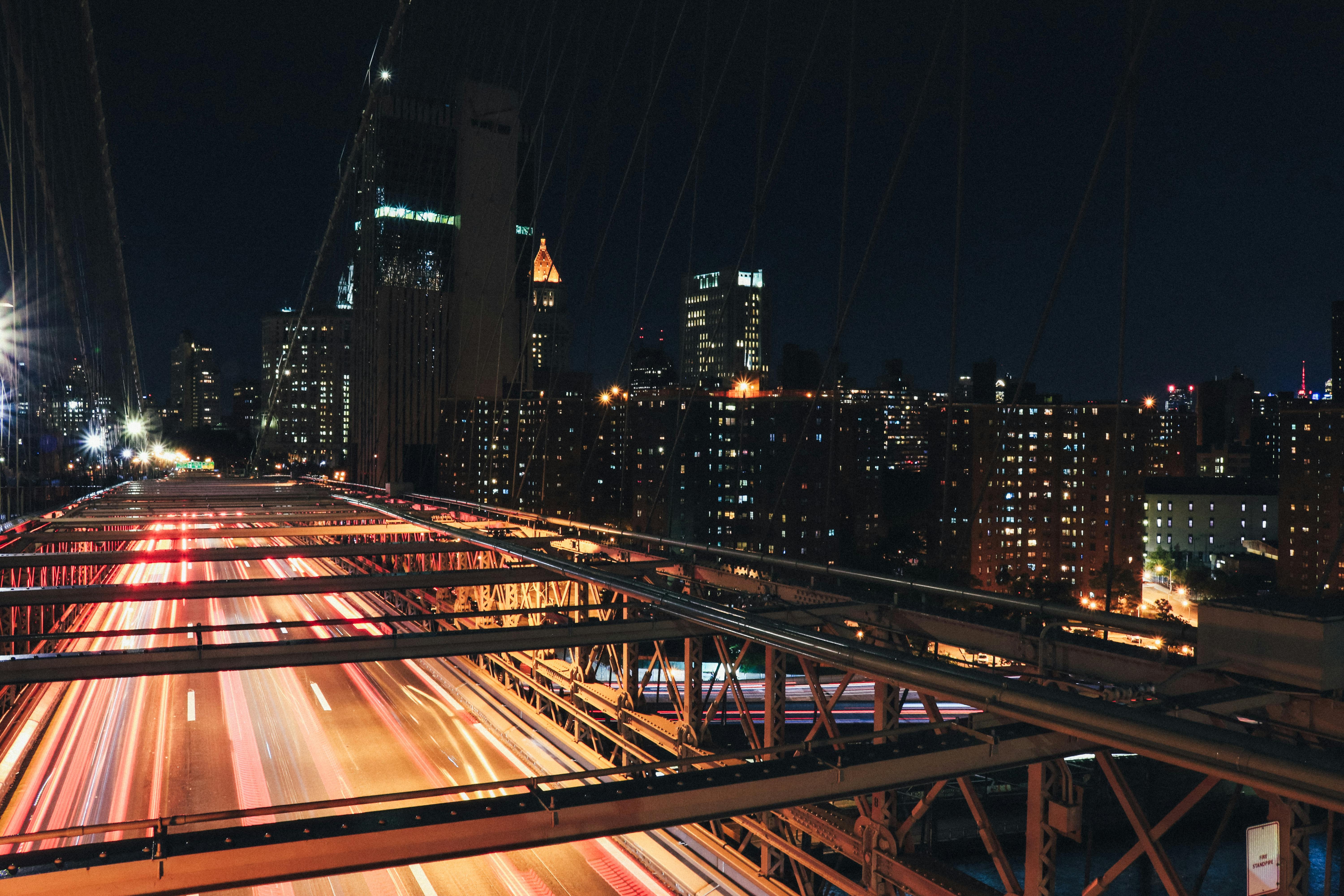 A long-exposure night shot from the Brooklyn Bridge, showing blurred light trails of traffic heading toward the lit-up New York City skyline.