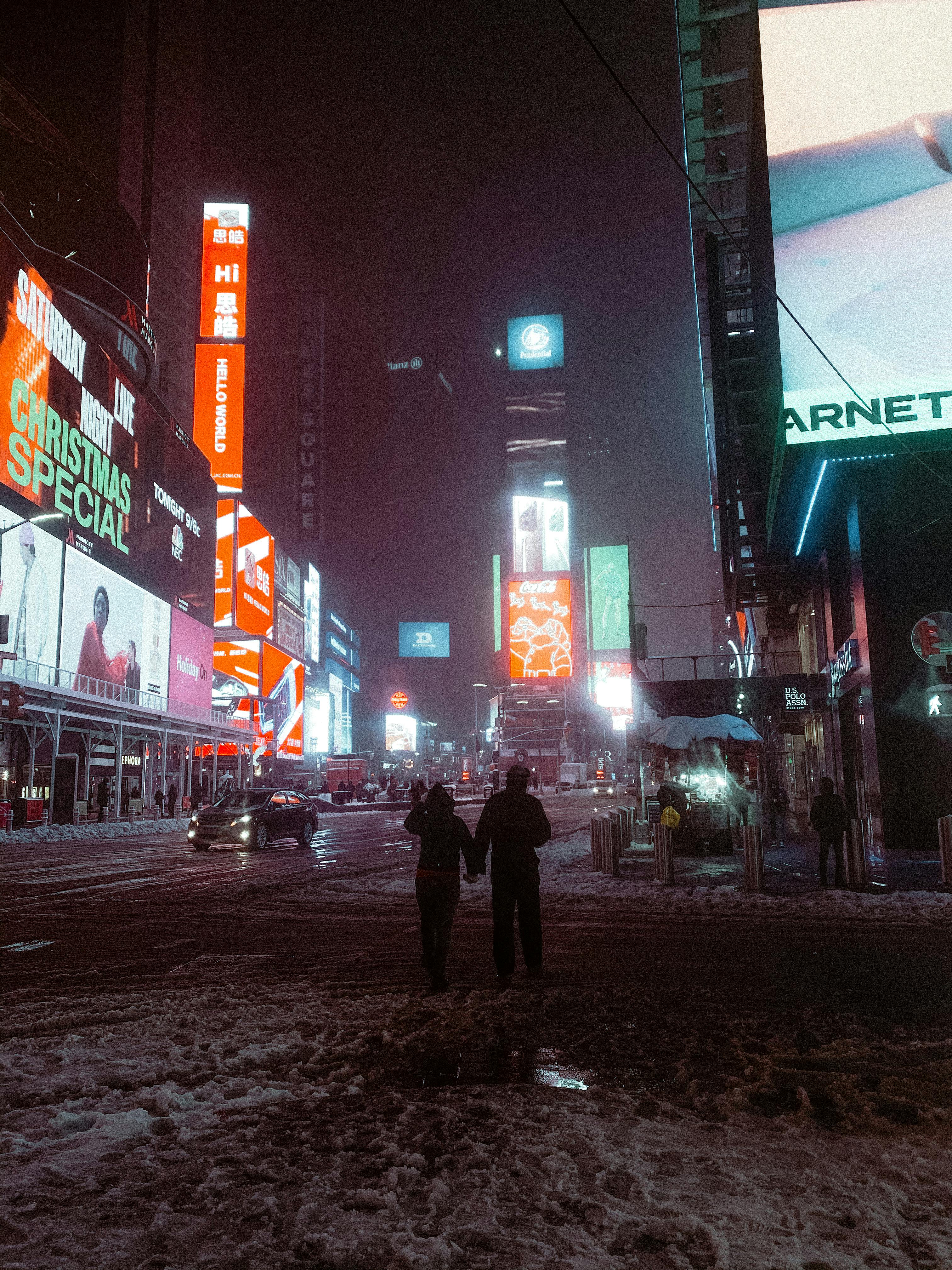 Snowy night in Times Square, New York, with the bright, colorful glow of digital billboards reflecting off the slushy street and a few pedestrians.